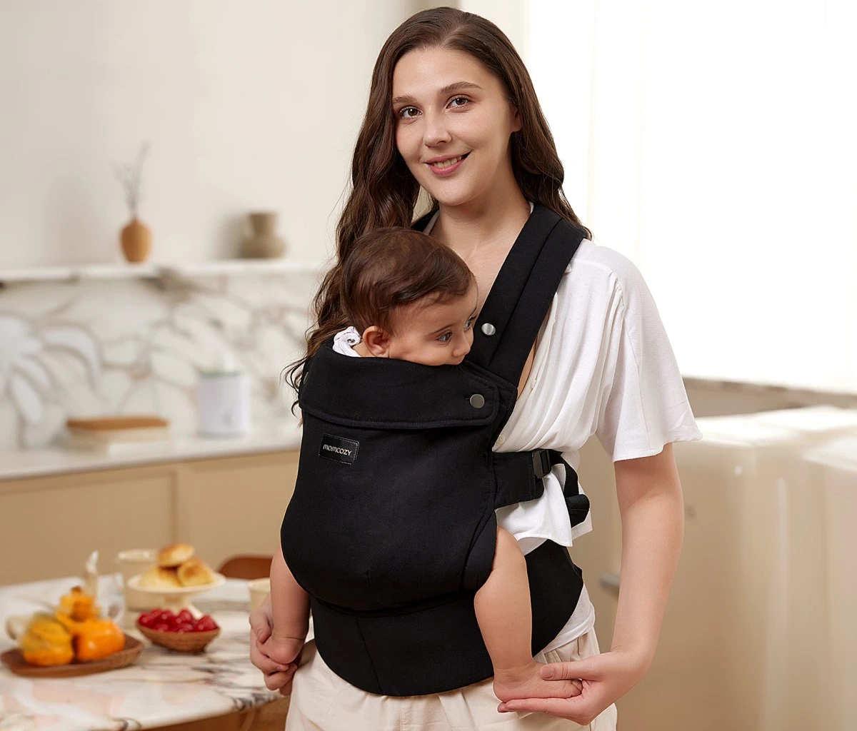 Uma mulher sorri enquanto segura um bebé num porta-bebés Momcozy preto numa cozinha luminosa