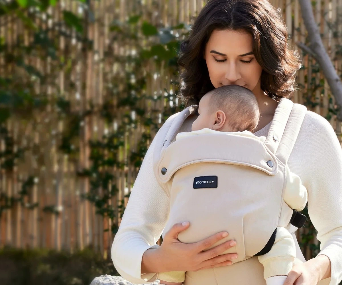 Uma mulher abraça um bebé num porta-bebés bege, com uma vedação de bambu ao fundo no jardim