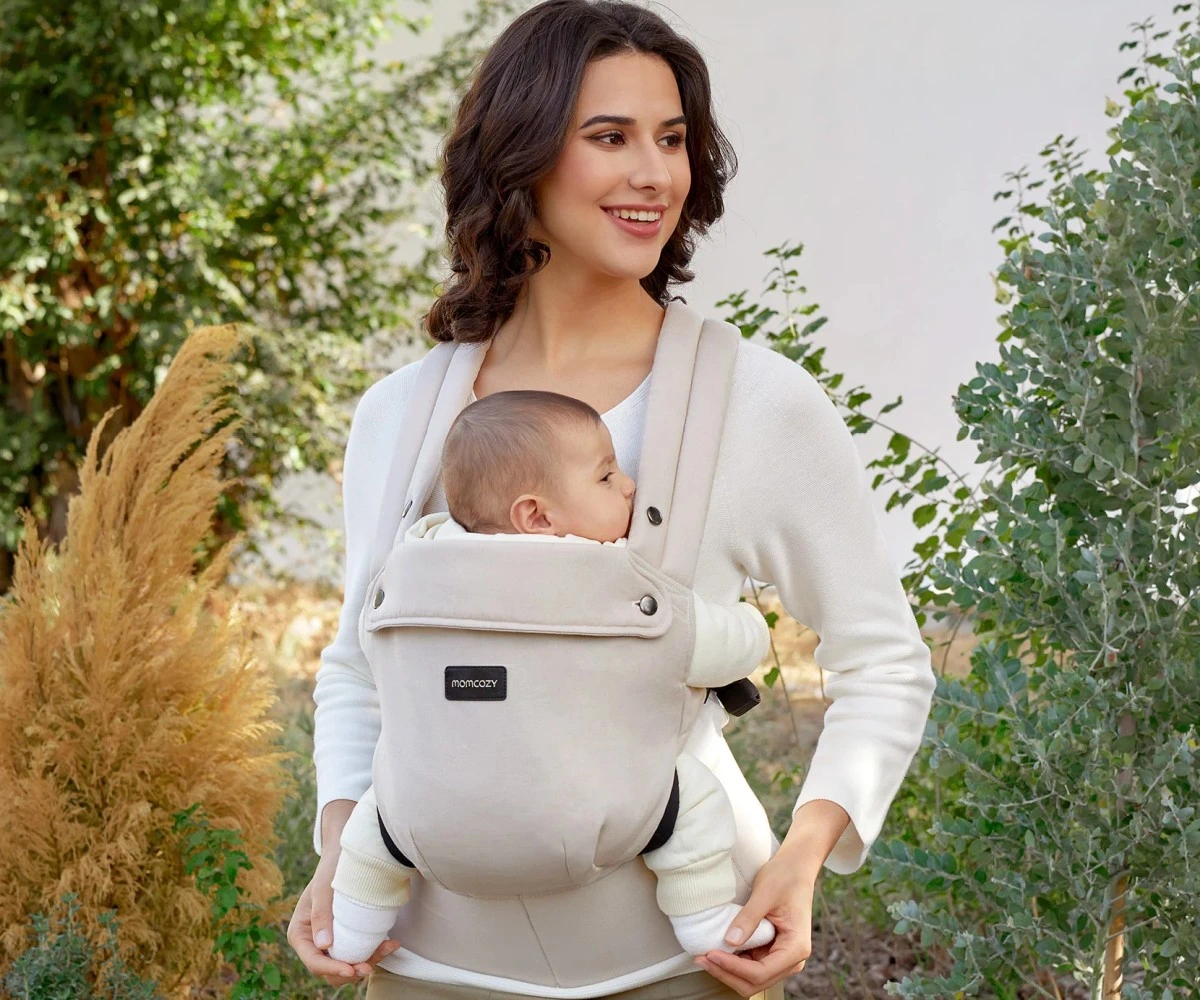 Mulher sorrindo, carregando um bebé num porta-bebés bege com arbustos verdes ao fundo